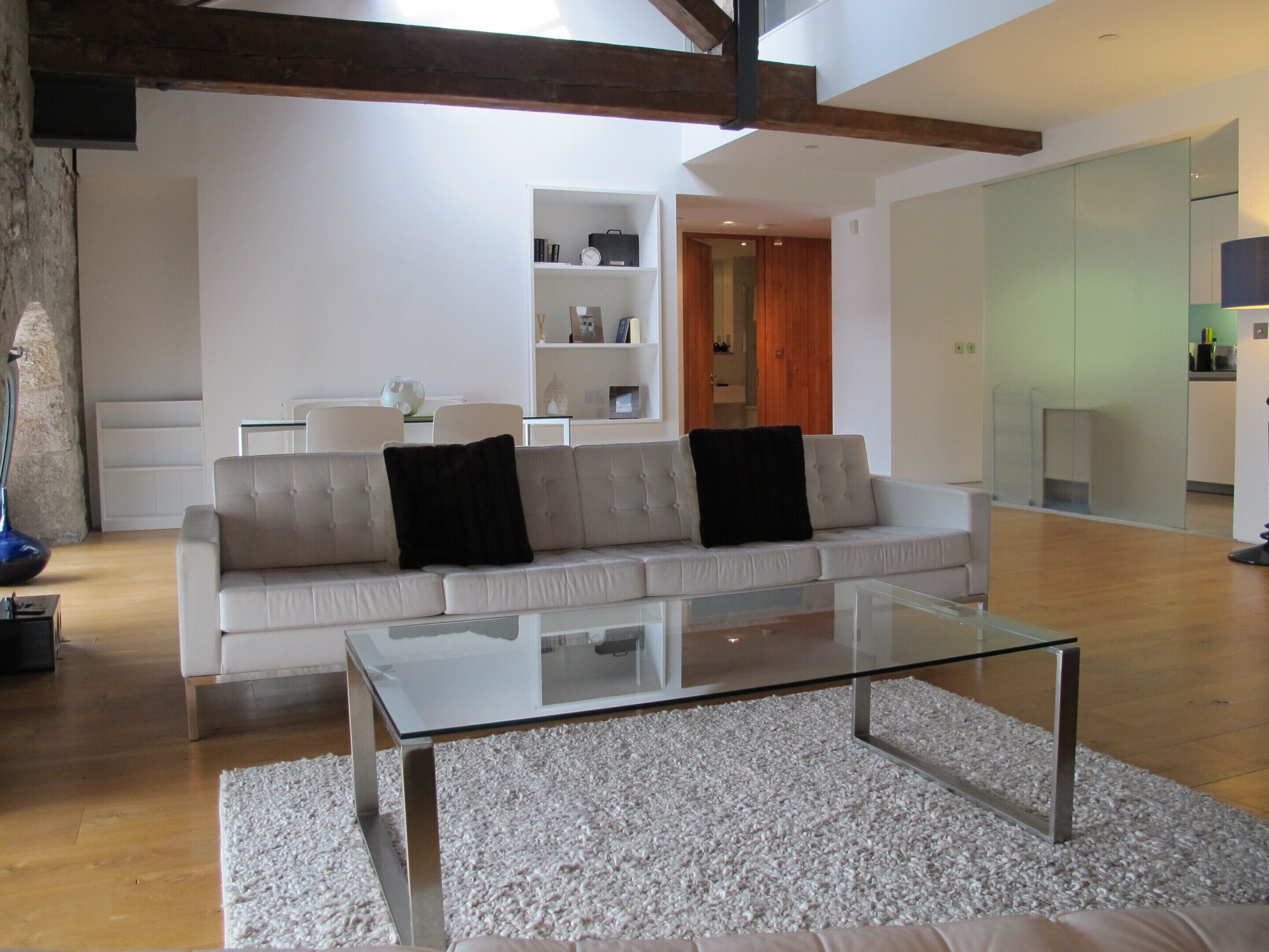 Una sala de estar moderna con una mesa de centro de vidrio, un sofá beige con cojines negros y una alfombra blanca.