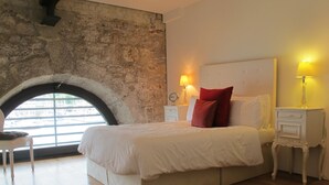 Un dormitorio con una cama grande, una pared de piedra, un ventanal y una mesita de noche con lámpara.
