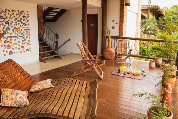 Una terraza de madera con un sofá, una silla de madera y una mesita. Hay plantas en macetas a lo largo de la barandilla.