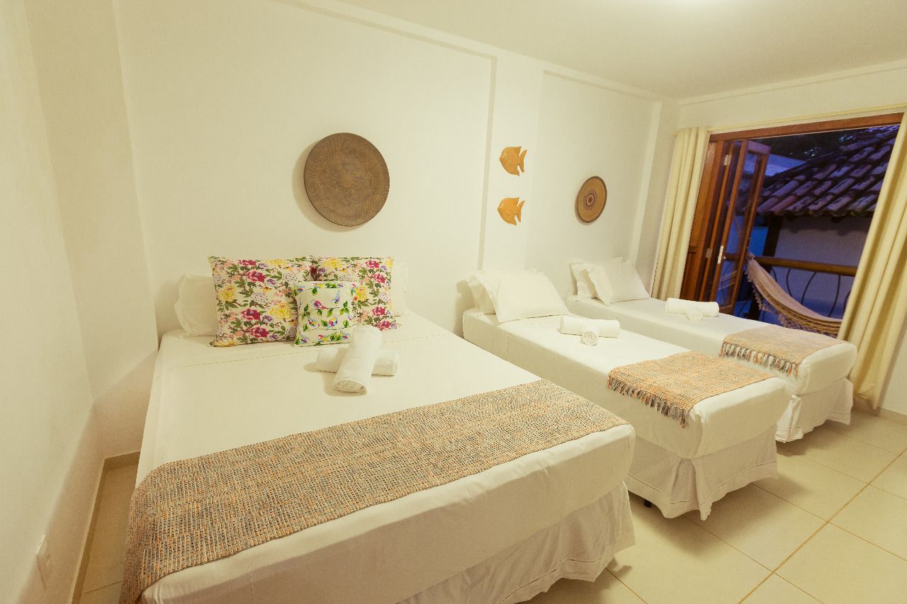 A hotel room with two beds, each with a floral pillow and a woven blanket. There are decorative wall plates and a view of the outside through a window with curtains.