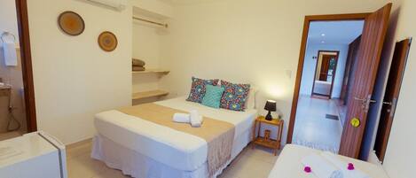 Quarto de hotel com uma cama, uma mesinha de cabeceira com abajur, uma pequena mesa com toalha e um frigorífico.
