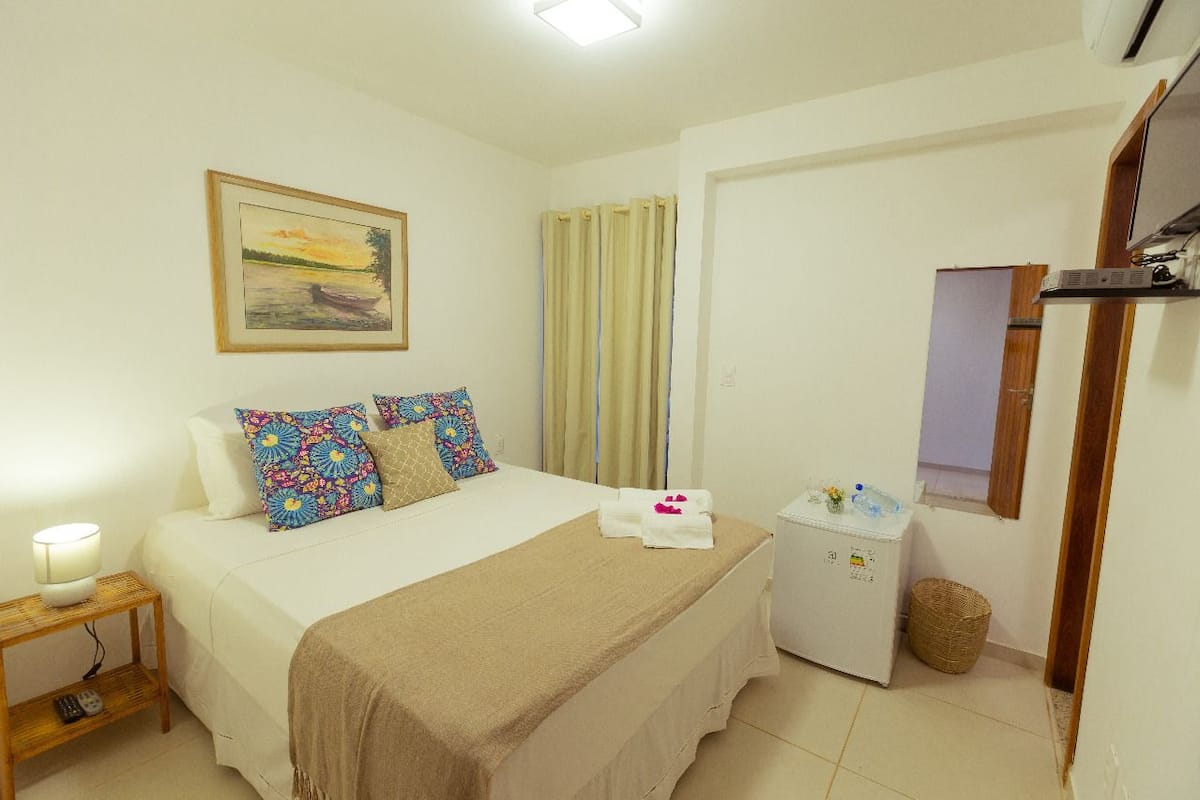 Quarto com cama, mesa de cabeceira com abajur, um pequeno frigorífico e janela com cortinas.