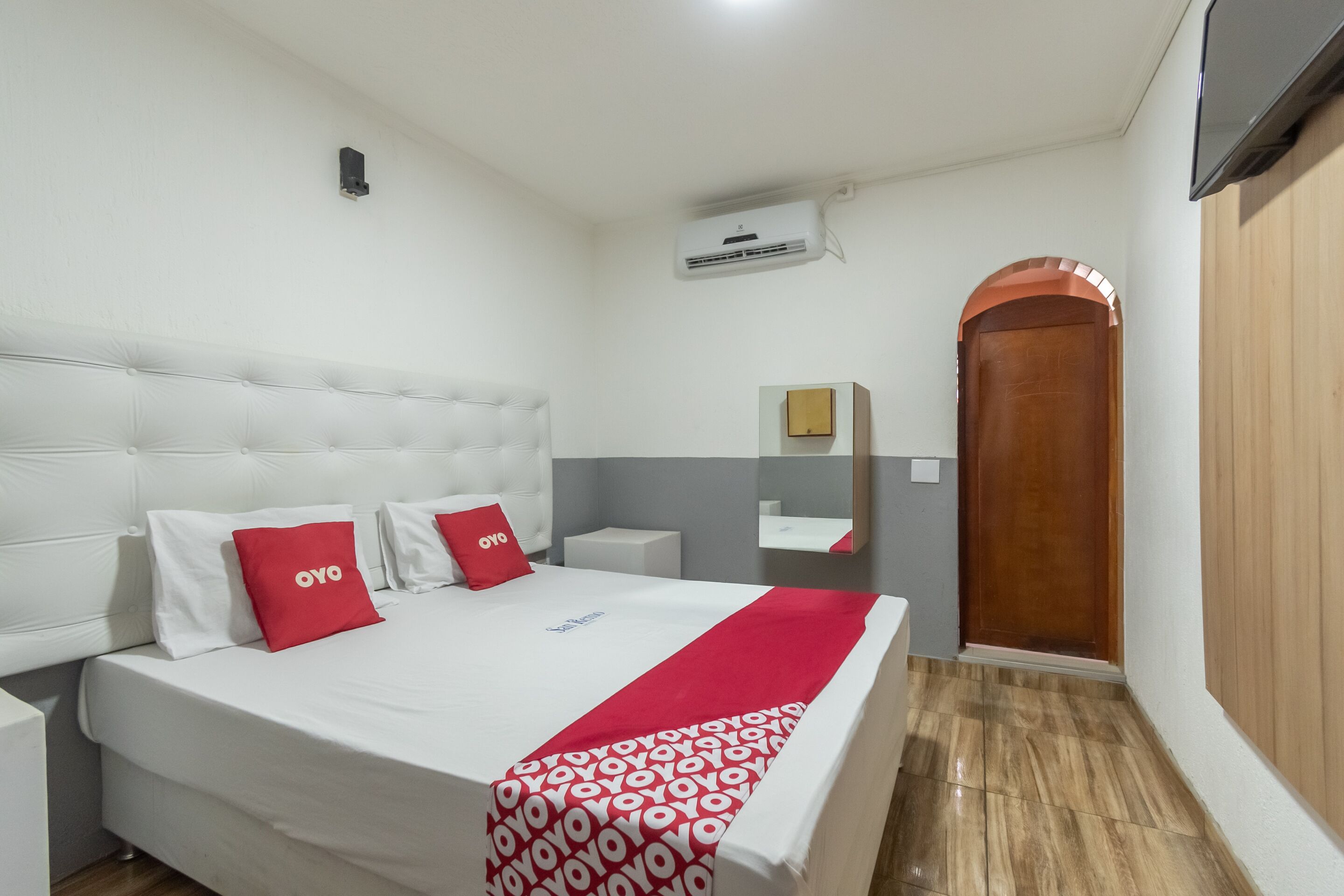 A hotel room with a bed, white walls, a wooden door, and a small bedside table.