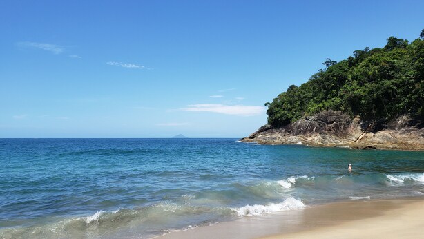 Ein Strand mit klarem, blauem Wasser, sandigem Ufer und einer bewaldeten Klippe.