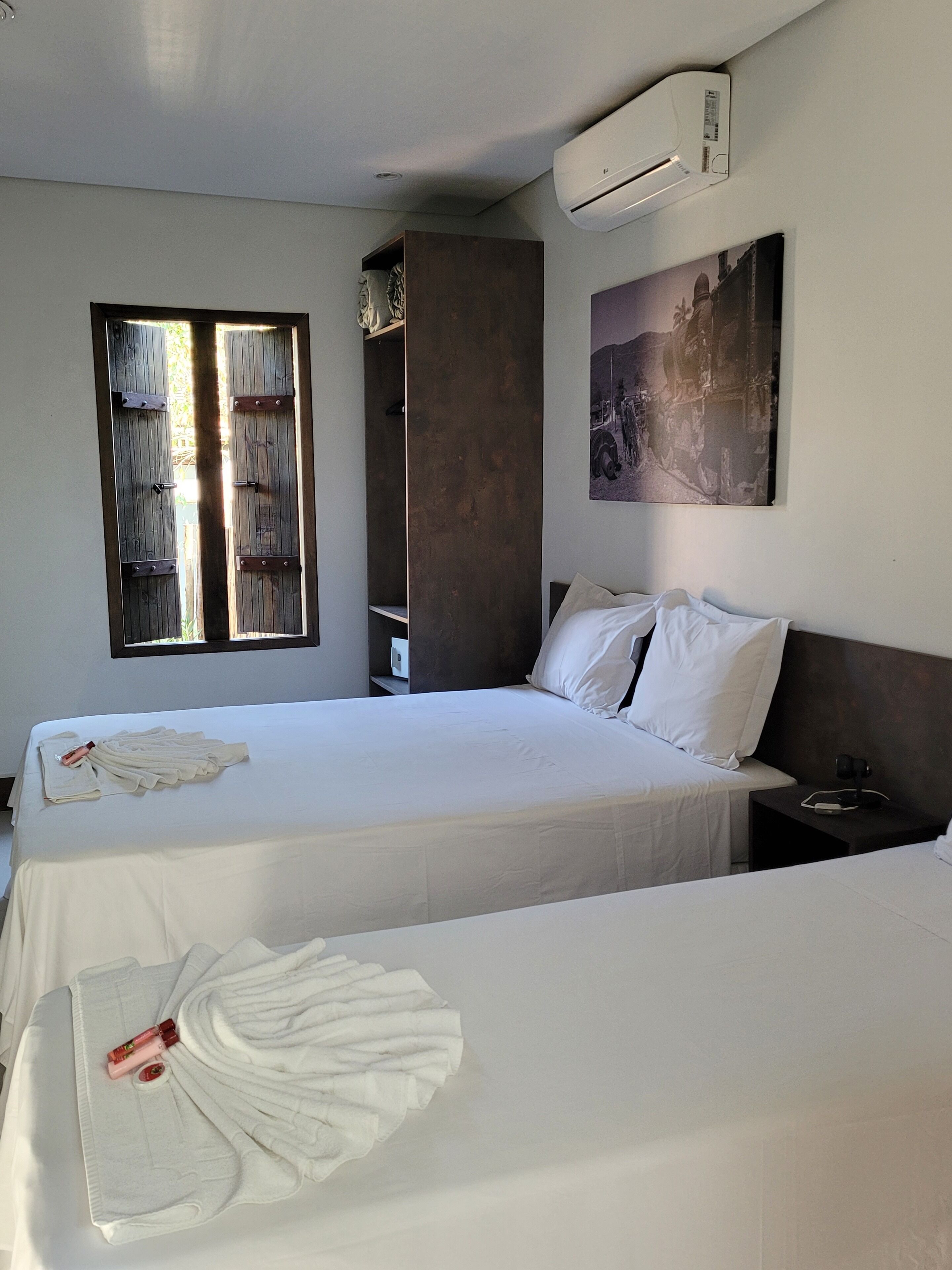 Una habitación de hotel con una cama grande, una cama más pequeña, un armario, una ventana y un aire acondicionado instalado en la pared.