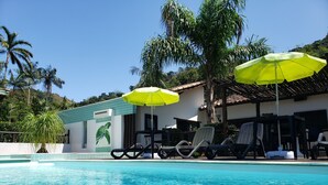 Ein Schwimmbecken mit klarem Wasser, umgeben von Liegestühlen und gelben Sonnenschirmen, mit einem Klinkergebäude und Palmen im Hintergrund.