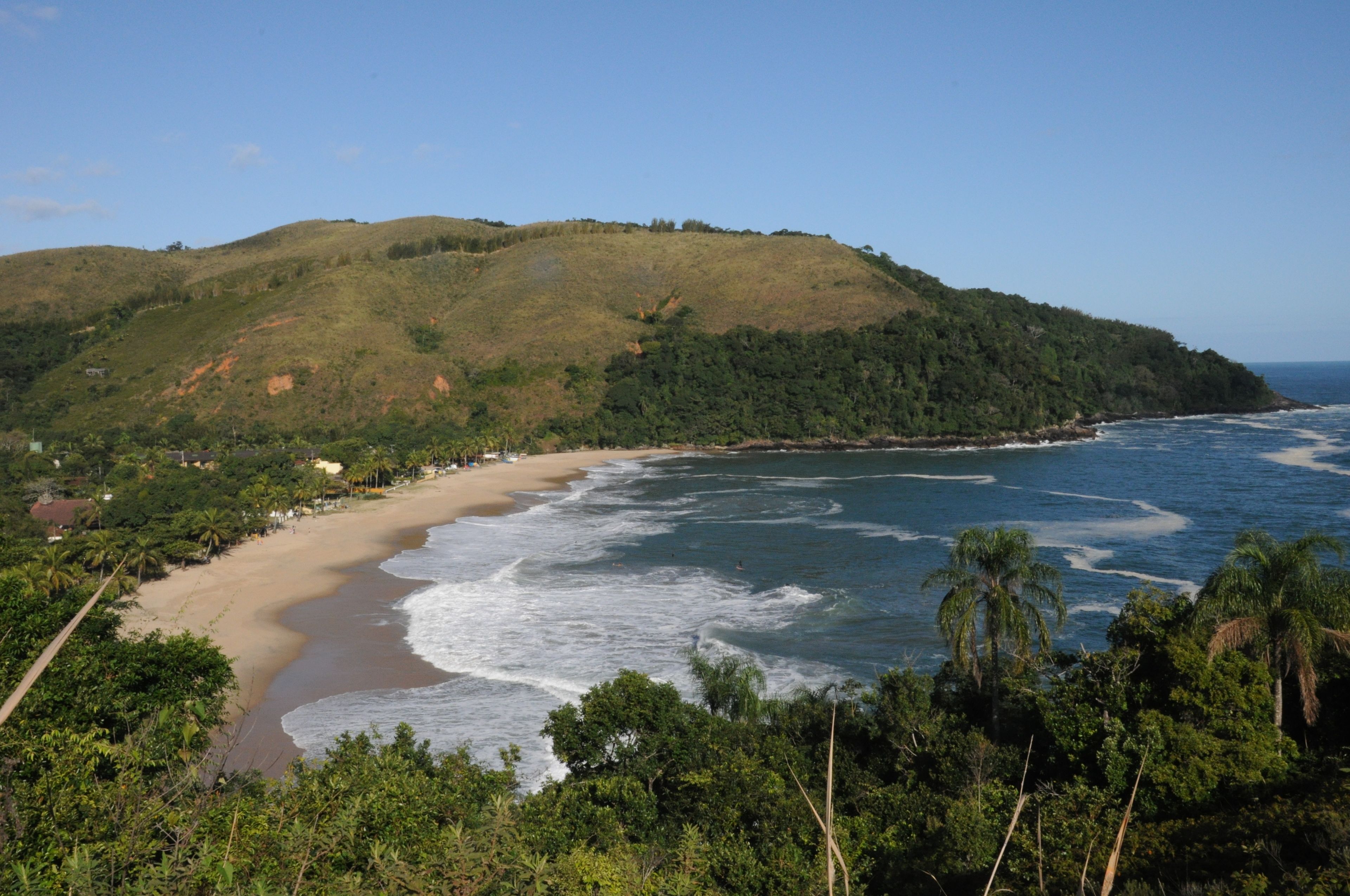 A coastal landscape with a beach, ocean waves, and a hill with dense vegetation.