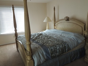 A four-poster bed with a floral bedspread, a bedside table with a lamp, and a window with blinds. - Windermere by the Sea #2A (Myrtle Beach)