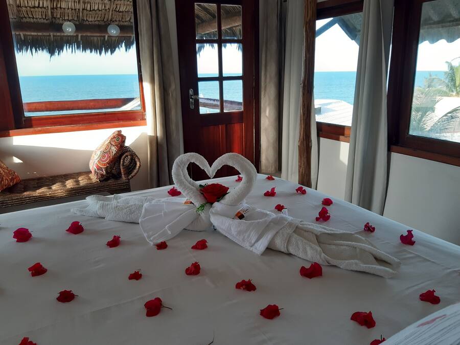 Una cama con una toalla dispuesta en forma de corazón y pétalos de rosa, una silla de mimbre y vistas al océano a través de una ventana.