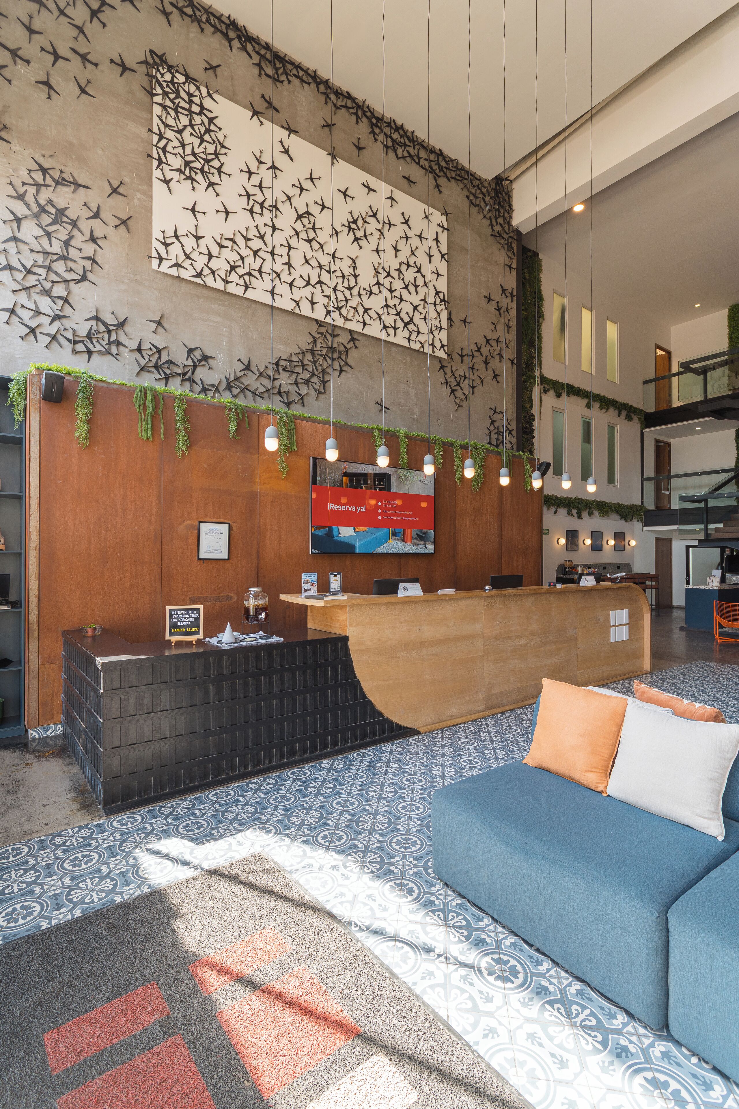 Ein Hotel-Empfangsbereich mit einem hölzernen Empfangstresen, einem blauen Sofa, einem gemusterten Teppich und einer Wand mit einem Vogel-Wandgemälde.