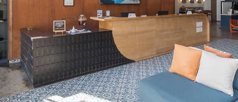 A hotel reception area with a wooden reception desk, a blue sofa, patterned carpet, and a wall with a bird mural.