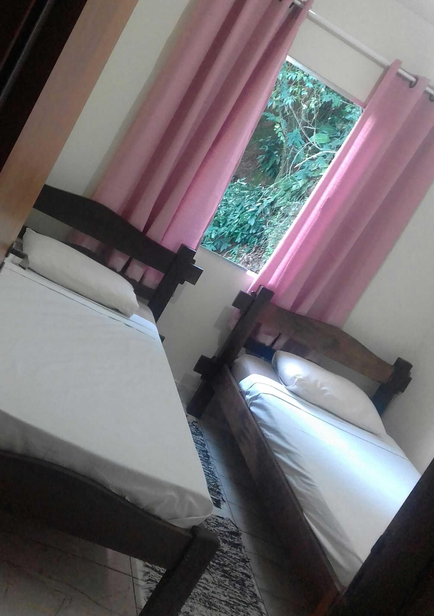Un dormitorio con dos camas individuales, una ventana con cortinas rosadas y vistas al verde exterior.