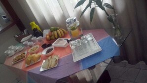 Una mesa de comedor dispuesta con diversos alimentos y condimentos, incluyendo pan, frutas y un dispensador de bebidas.