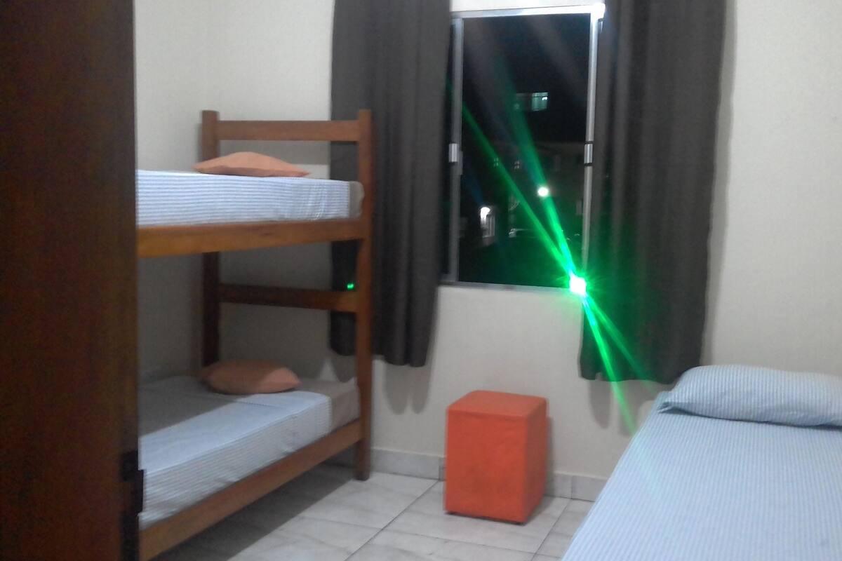 A small room with bunk beds, a single bed, a window with curtains, and a small orange stool.