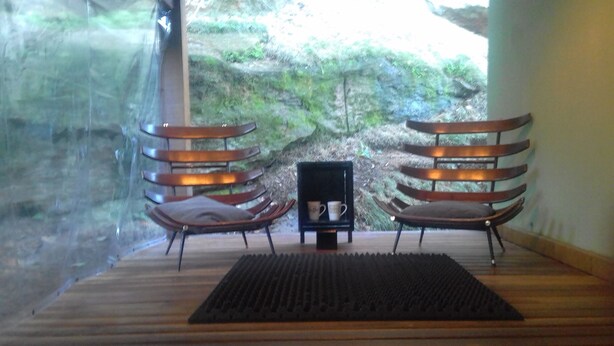 Una habitación con dos sillas de madera, una mesita con dos tazas y vistas a un terreno rocoso a través de una ventana.