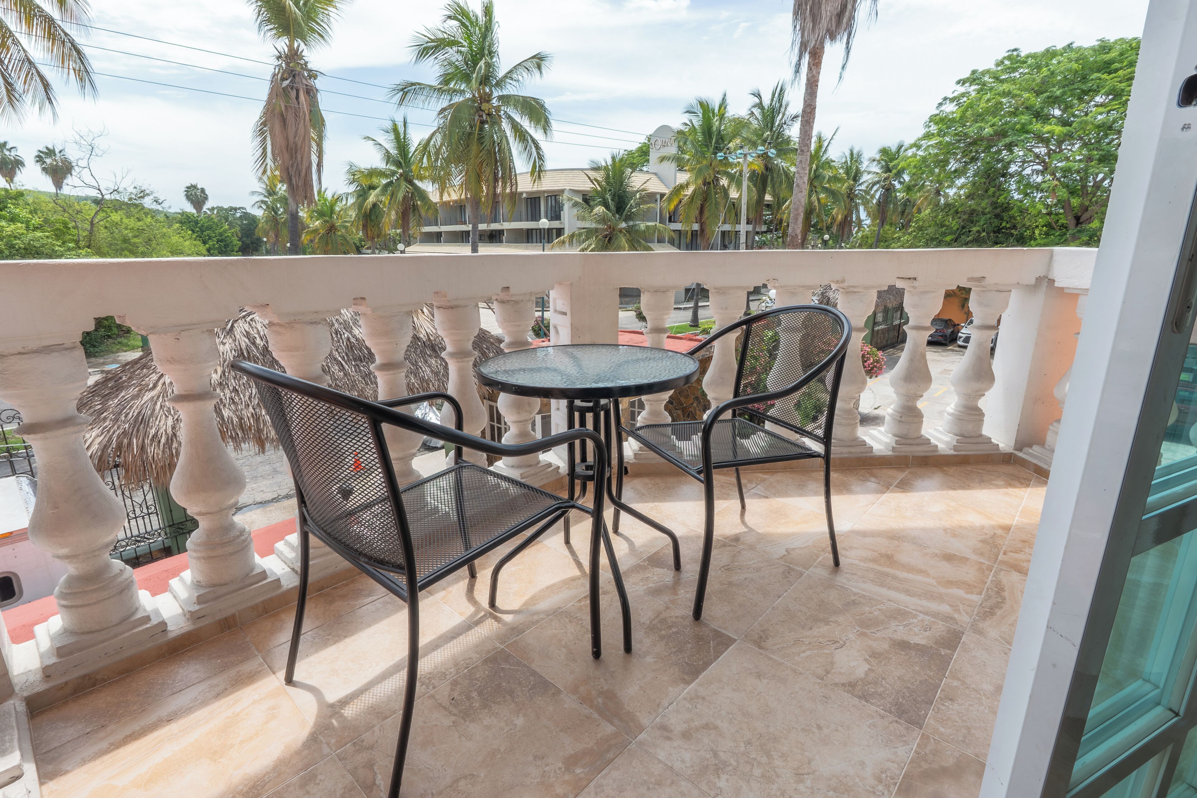 Ein Balkon mit Tisch und zwei Stühlen, einer Steinsichtschutzwand und Palmen im Hintergrund.