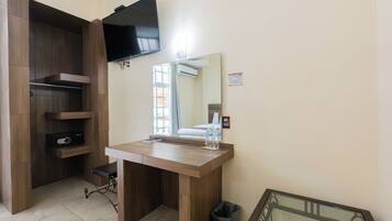 A hotel room with a wooden desk, a television mounted on the wall, a window with curtains, and a trash bin.