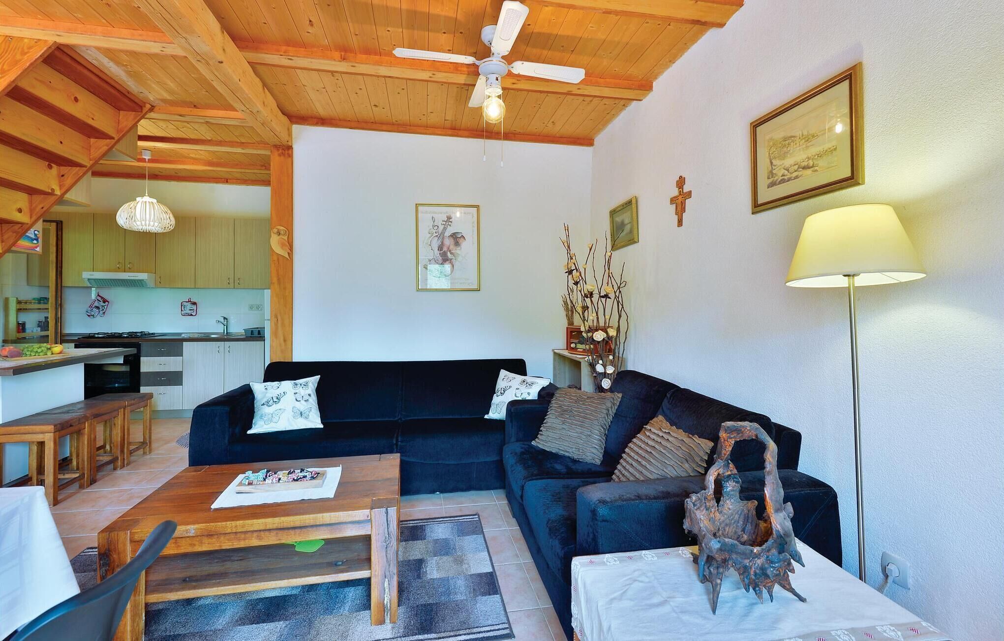 A cozy living room with a dark blue sofa, wooden coffee table, and a lamp. A kitchen is visible in the background with wooden cabinets and a hanging light fixture.