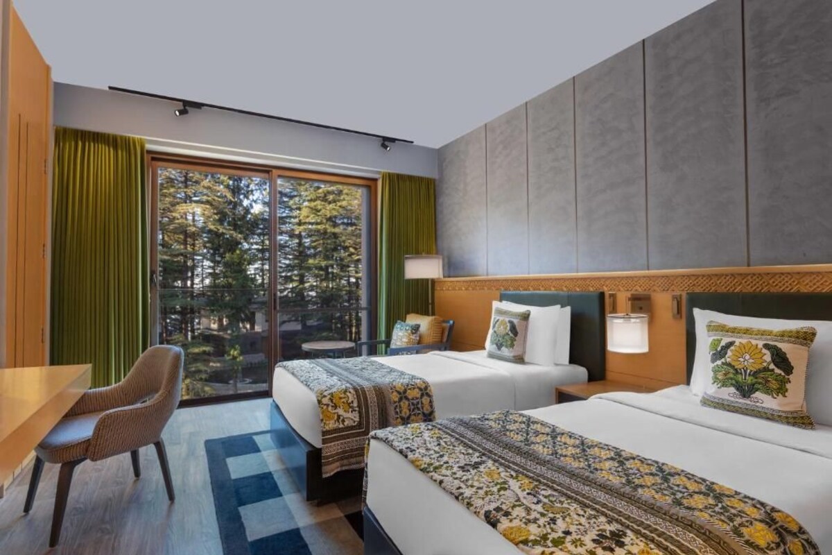 A hotel room with two beds, a desk, and a view of trees through the window.