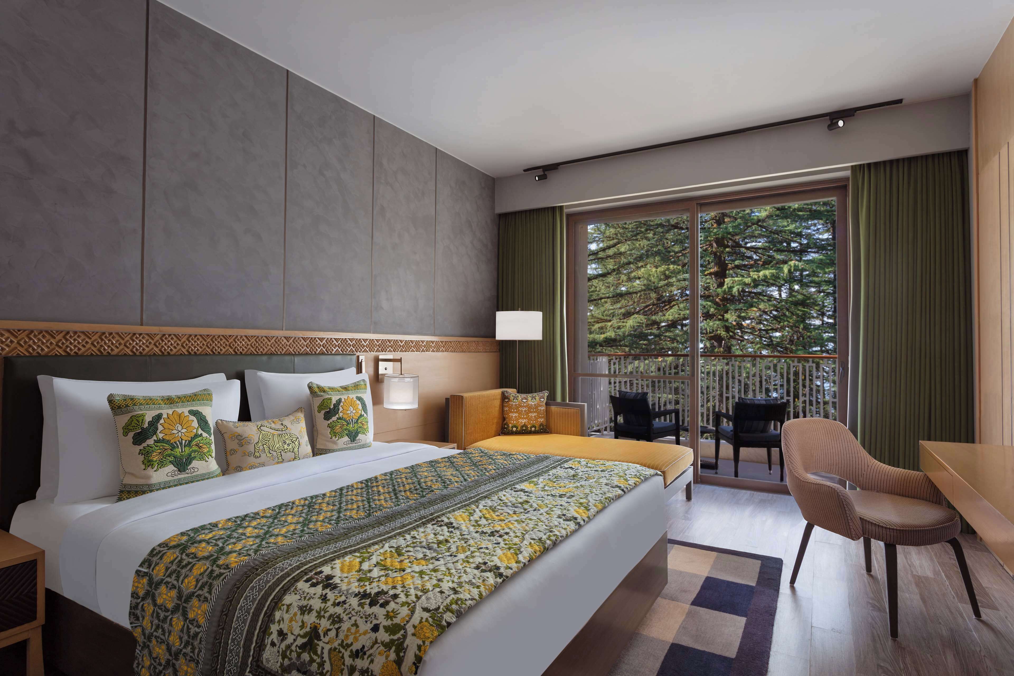 A hotel room with a large bed, a balcony with a view of trees, and a seating area with a chair and table.