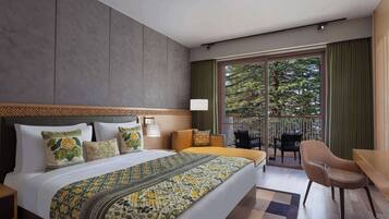 A hotel room with a large bed, a balcony with a view of trees, and a seating area with a chair and table.