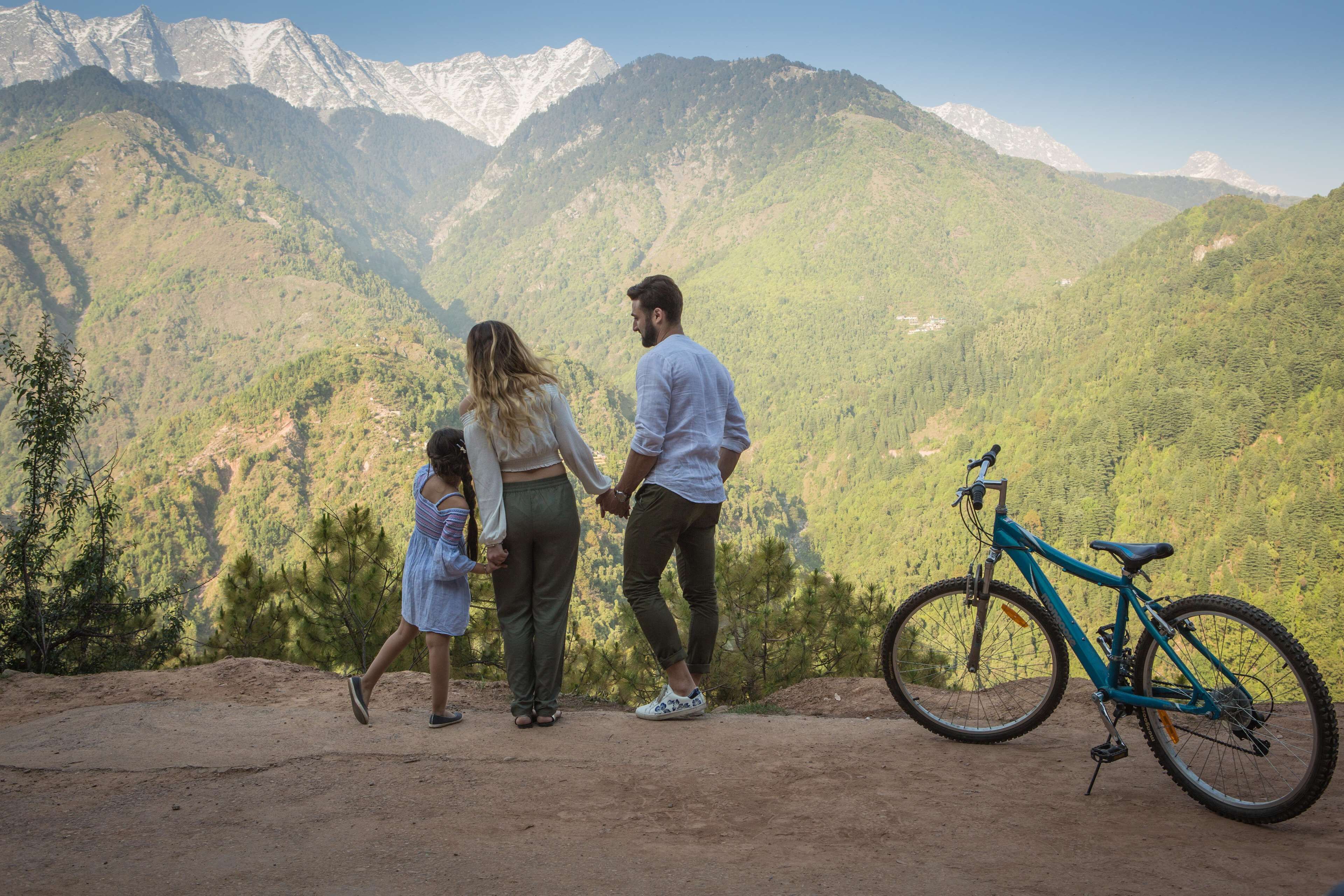 A family standing on a dirt path with a mountain view, holding hands, and a bicycle parked nearby.