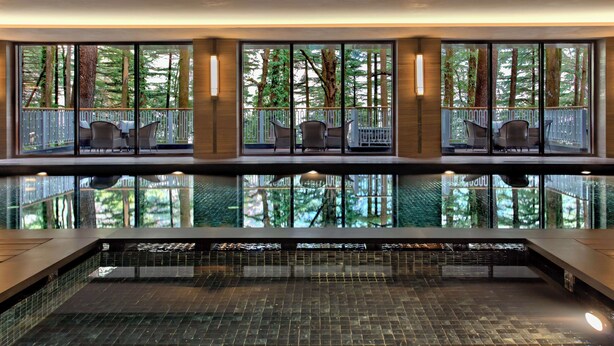 A modern indoor swimming pool with clear glass walls, multiple seating areas, and a view of a forested area. - Hyatt Regency Dharamshala Resort (Dharamshala)