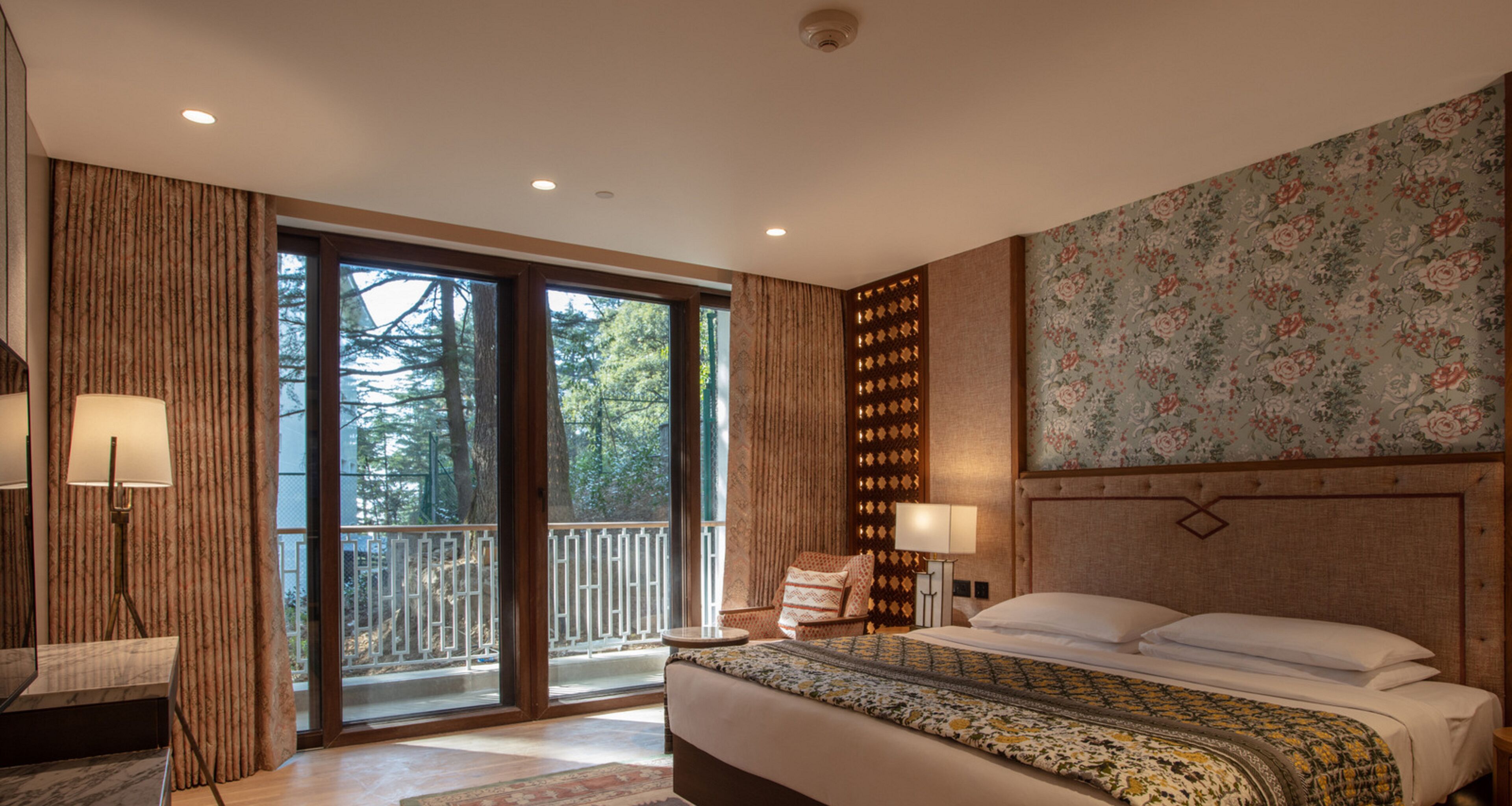 A hotel room with a large bed, a nightstand, a lamp, a view through the glass doors, and a floral wallpaper.
