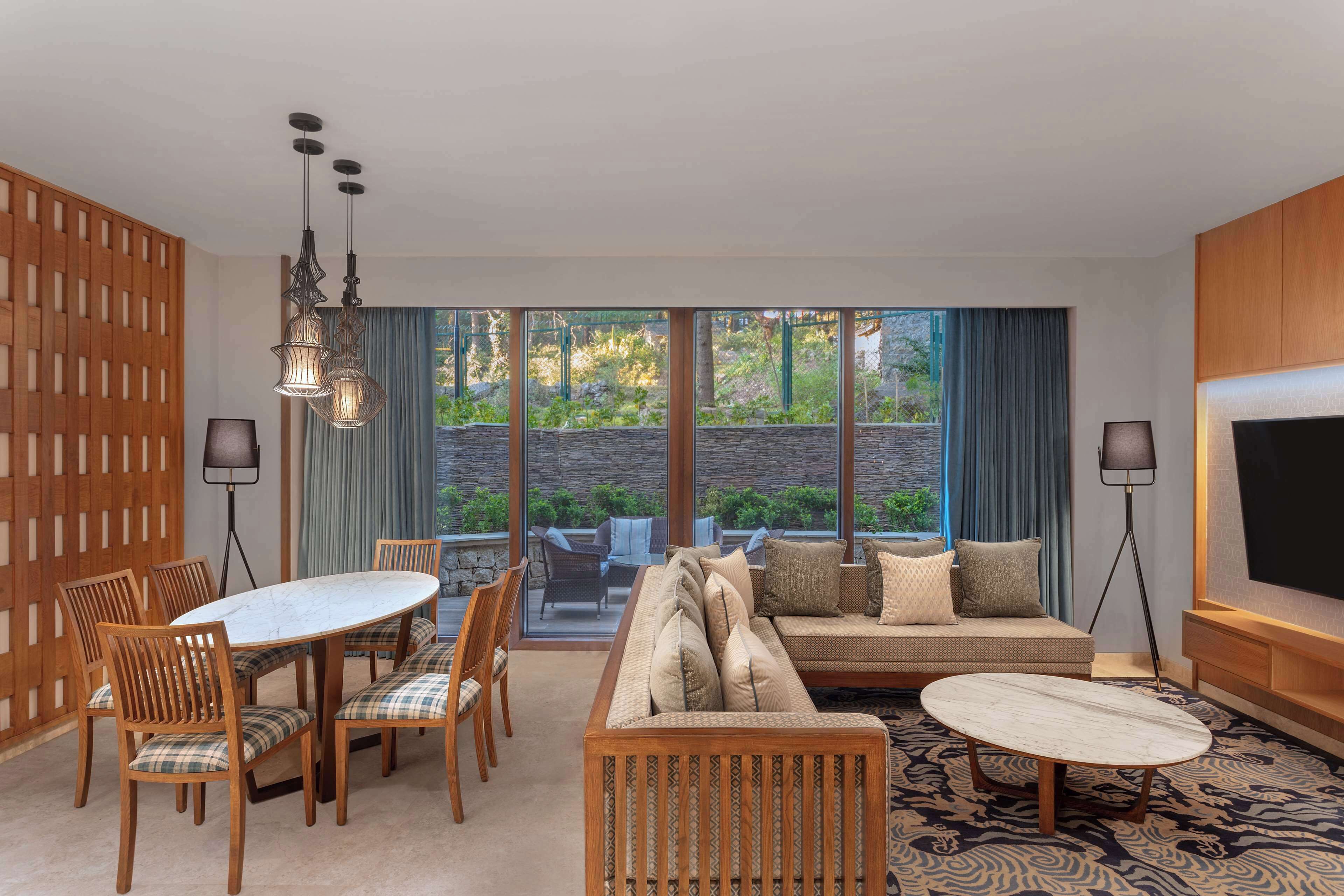 A hotel room with a dining area, a sofa, a TV, and large windows with a view of greenery.