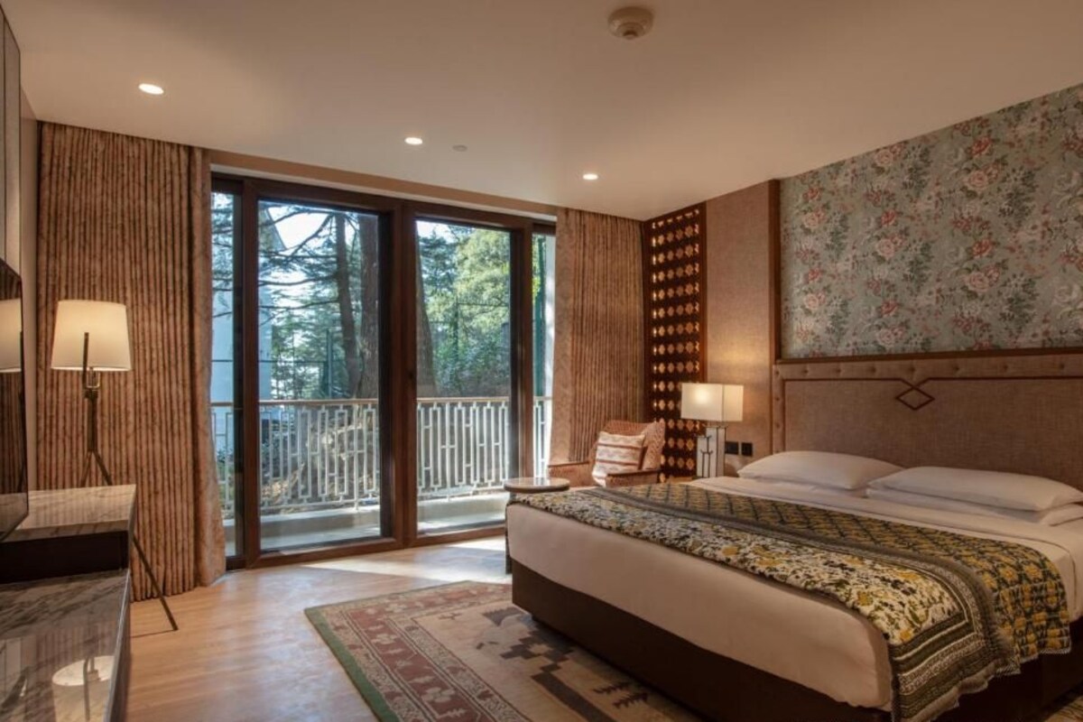 A bedroom with a large bed, a desk, a chair, a lamp, and a view of the outdoors.