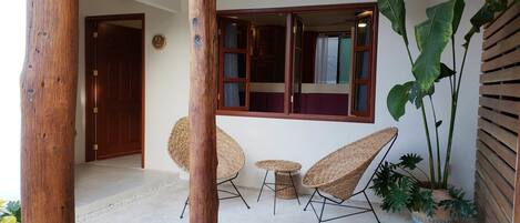 A room with a wooden door, a window with curtains, two wicker chairs, a small round table, and a potted plant.