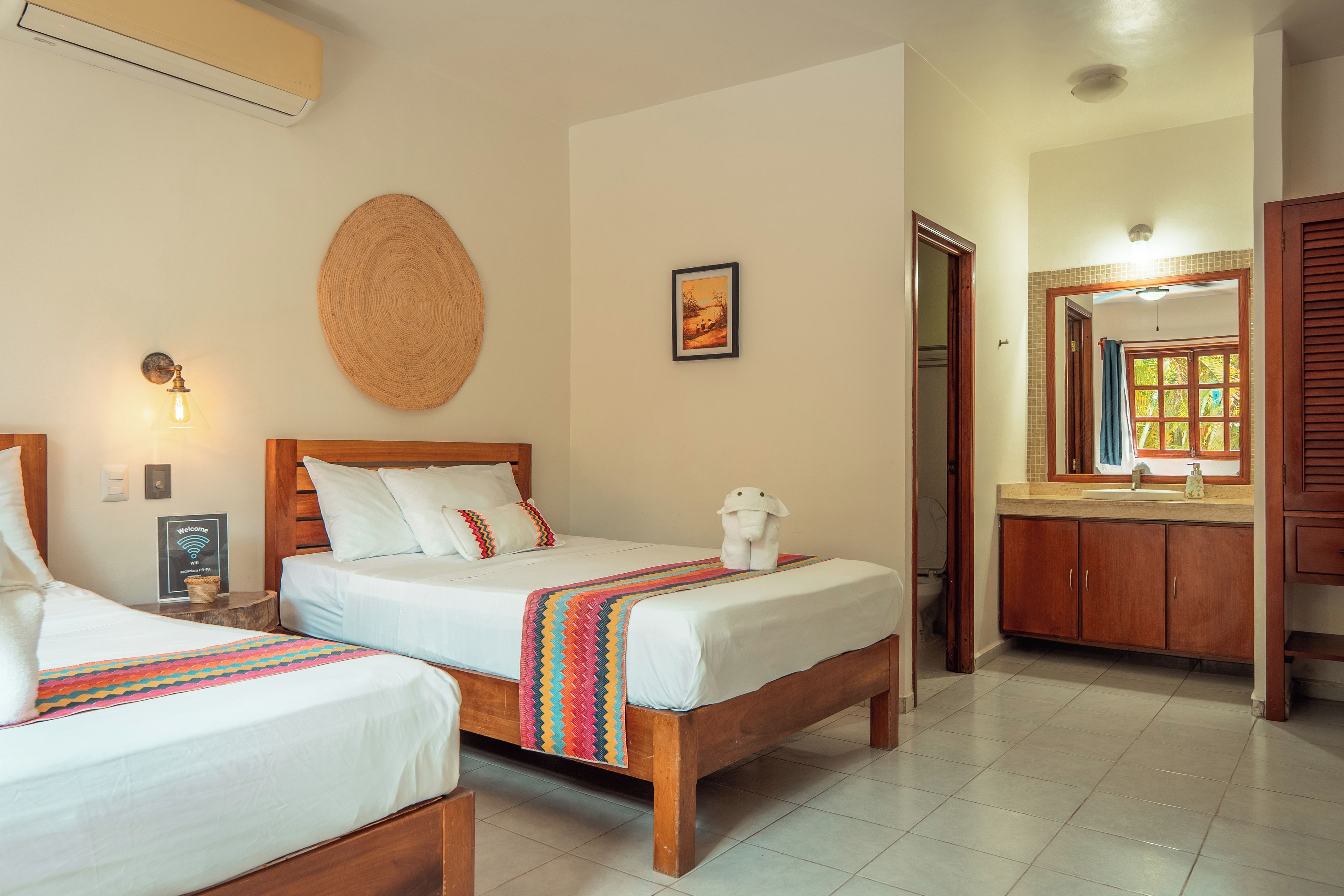 A hotel room with two wooden beds, a woven wall hanging, a framed picture, and a bathroom visible through an open door.