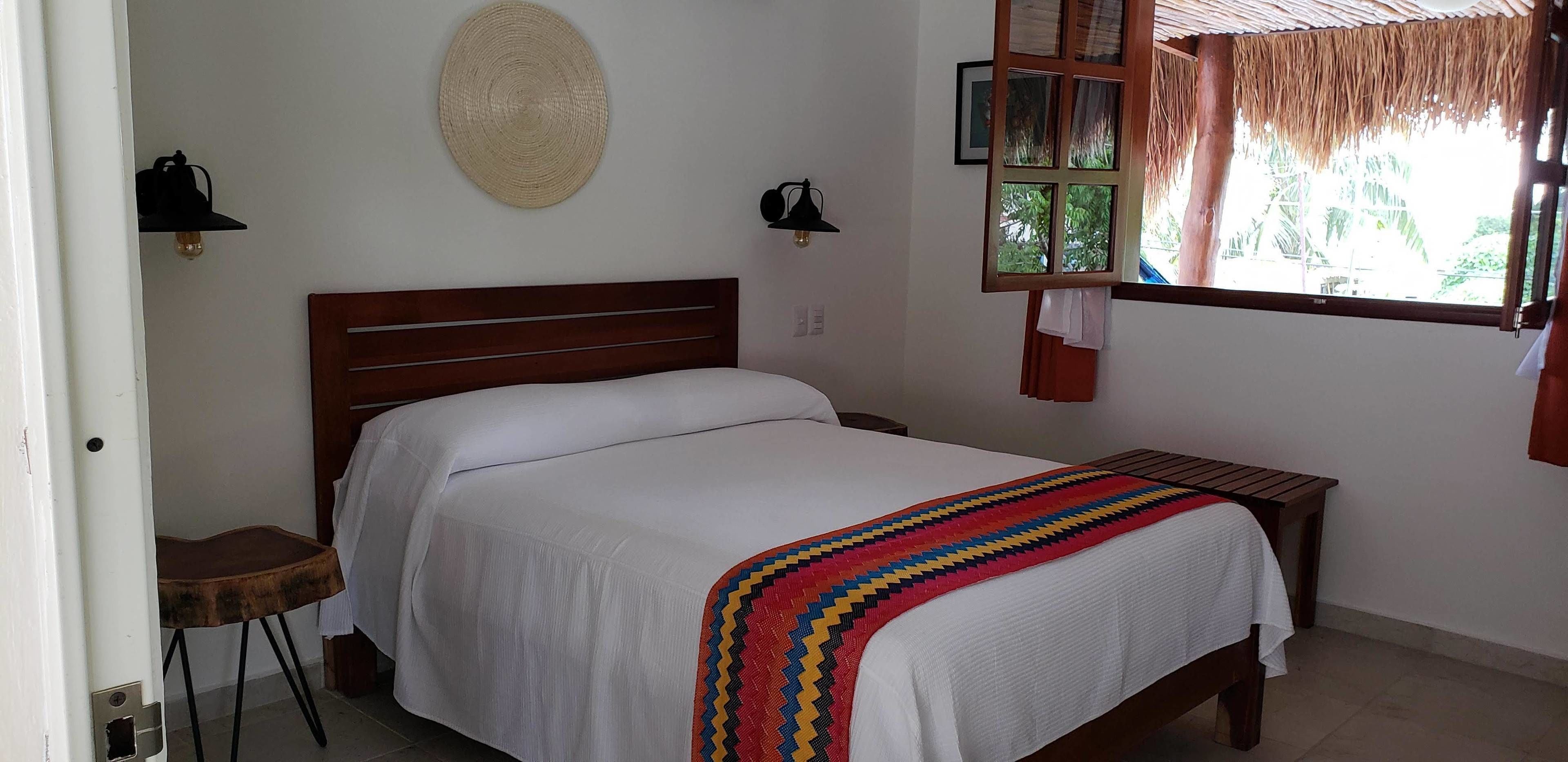 A neatly made bed with a white sheet and a colorful blanket, a wooden nightstand with a lamp, a woven wall hanging, and a window with a view of greenery.