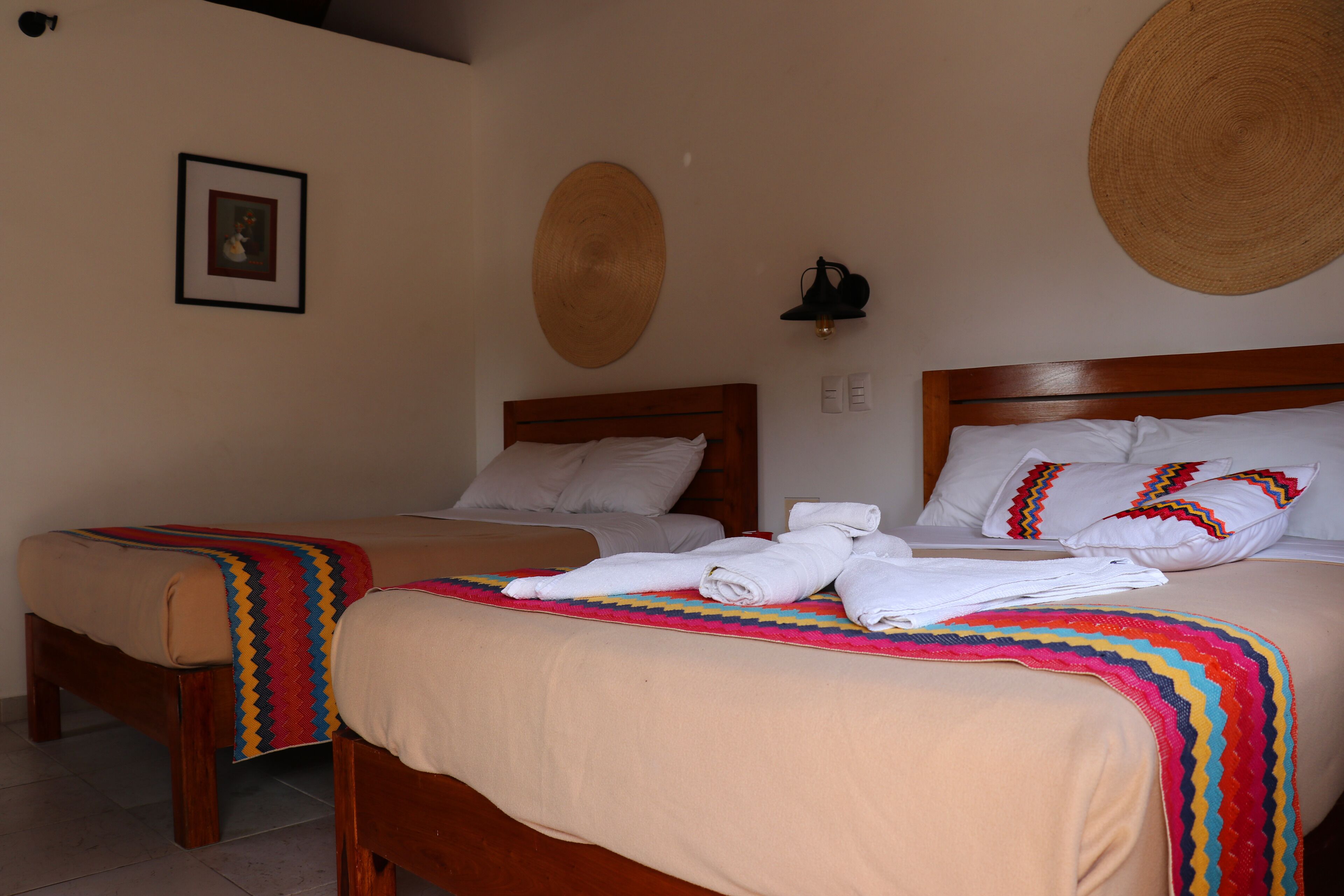 Una habitación de hotel con dos camas, cabecero de madera, manta de colores y un tapiz tejido en la pared.