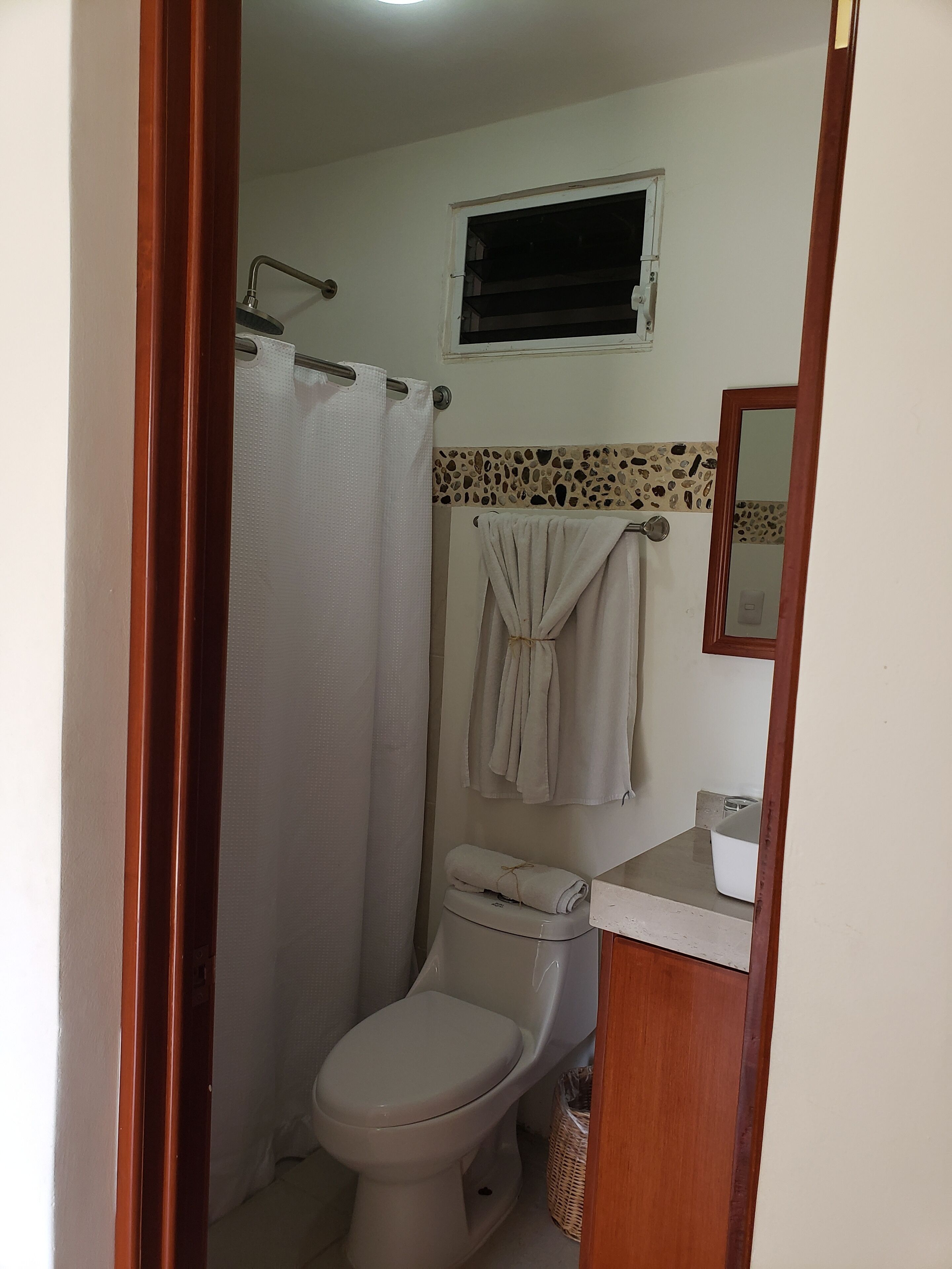 A bathroom with a toilet, a shower curtain, a towel rack with towels, a small window, and a mirror.