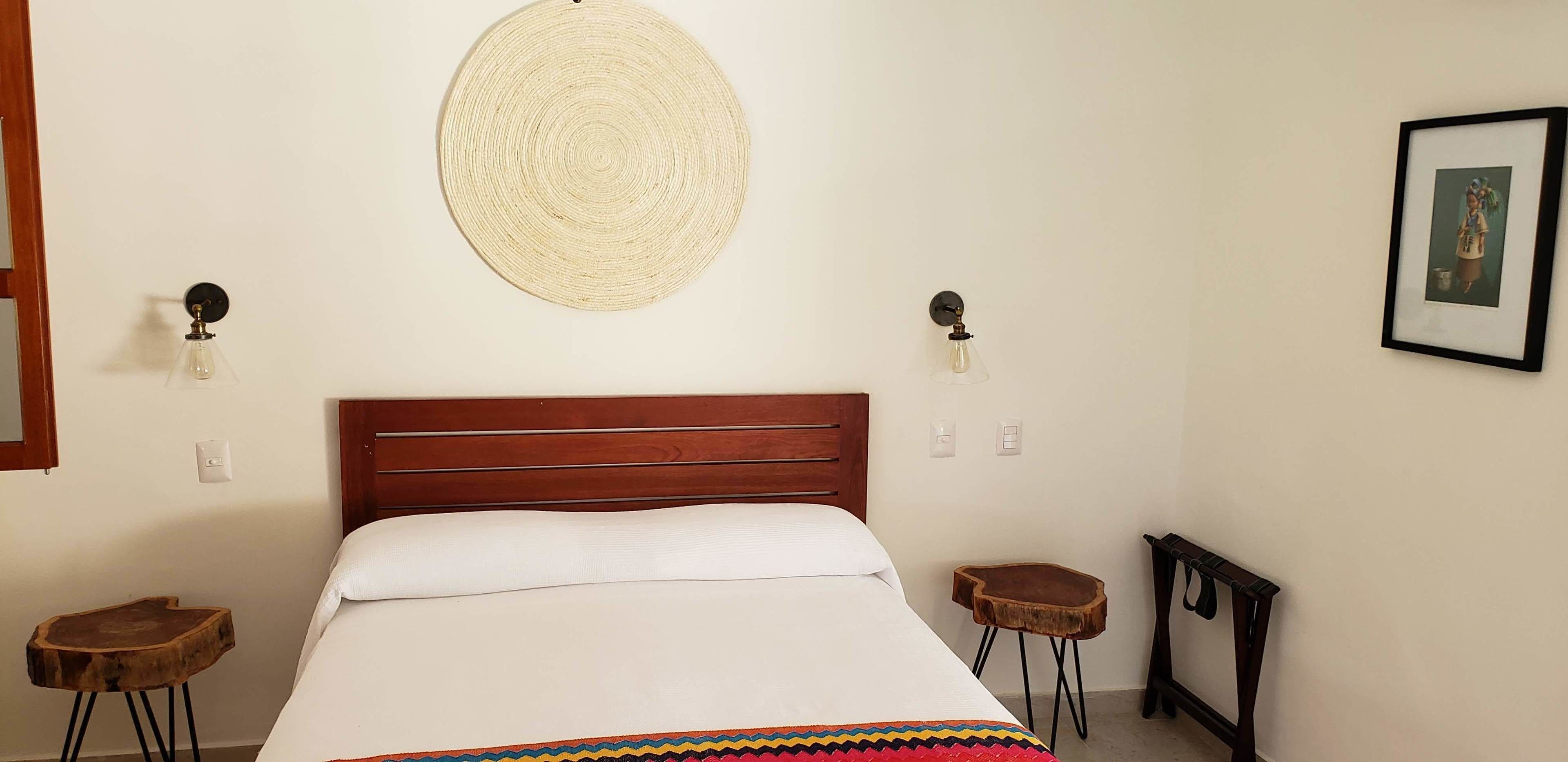 Un dormitorio con cabecera de madera, un tapiz redondo tejido en la pared, dos lámparas fijadas a la pared, un cuadro enmarcado y un taburete pequeño de madera.
