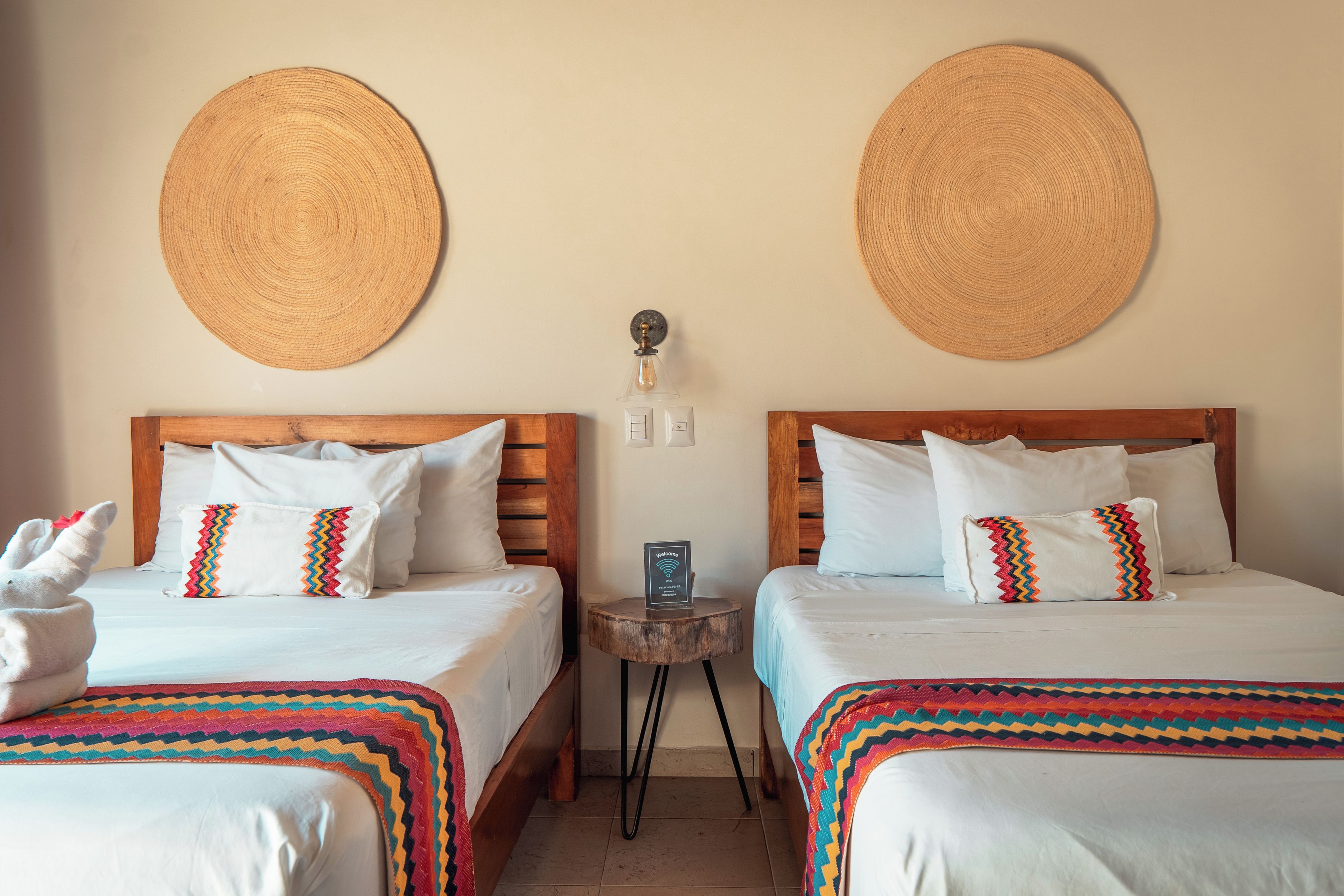 A hotel room with two beds, each with a wooden headboard, white bedding, and colorful pillows. There are woven wall hangings above the beds and a small table with a lamp between them.