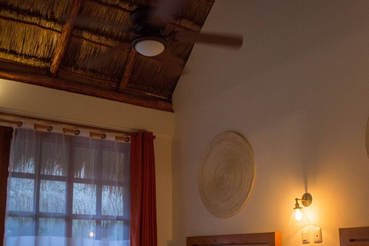 Ett rum med två sängar, en takfläkt, ett fönster med gardiner och en väggmonterad lampa.