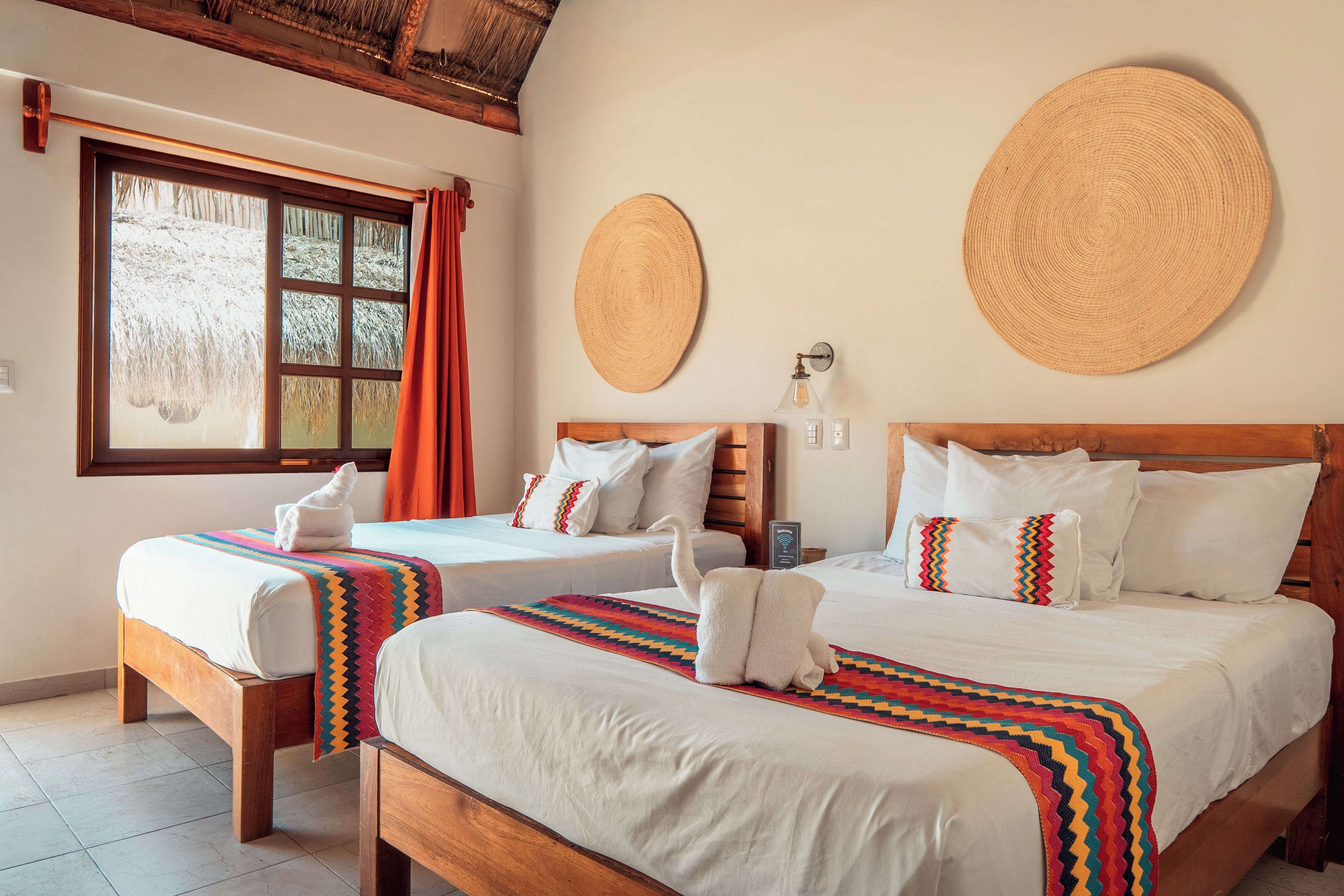 A hotel room with two beds, a window with curtains, a wall-mounted lamp, and a welcome sign.