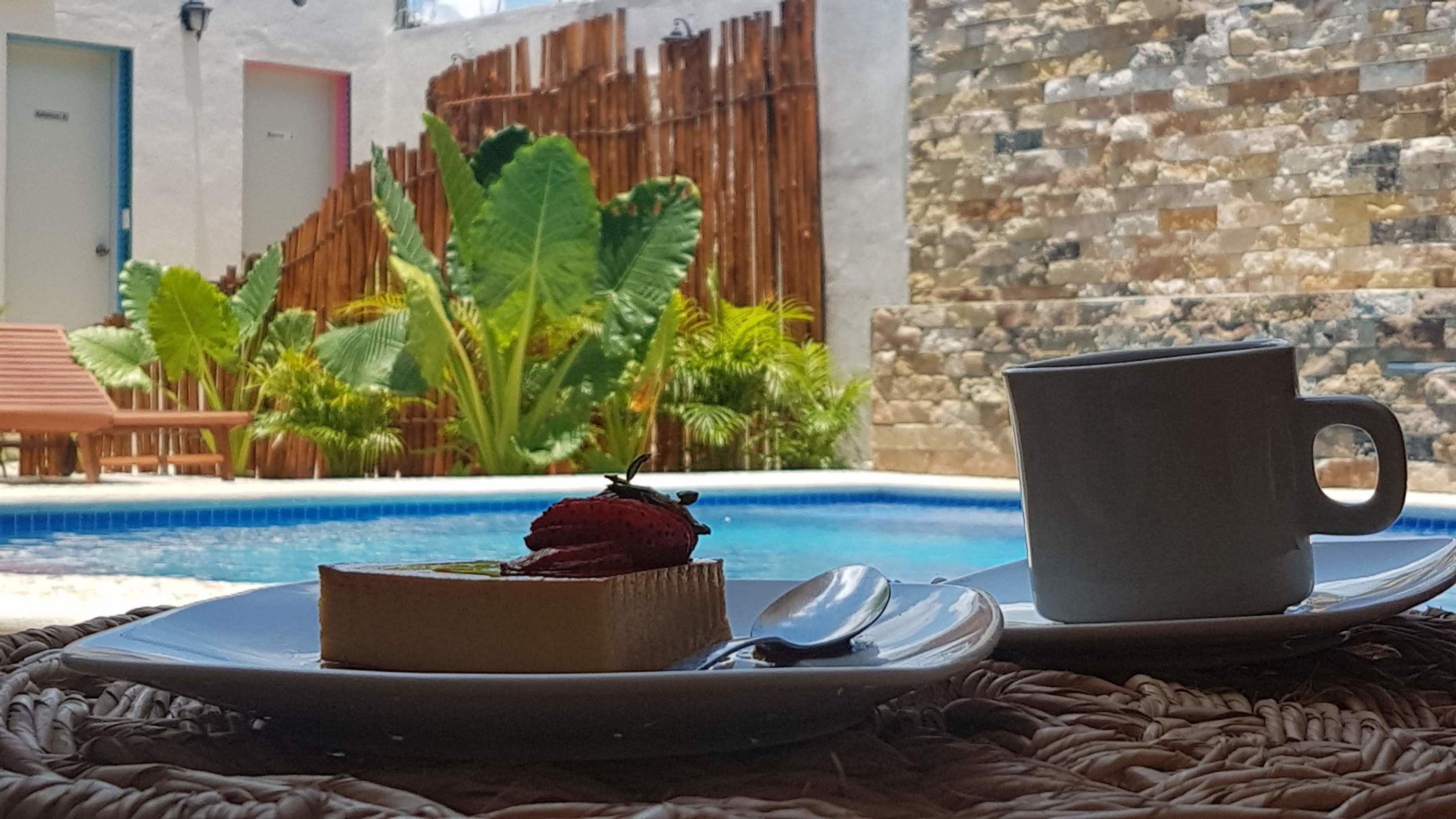 A dessert on a plate with a spoon and a cup on a woven mat by a poolside.