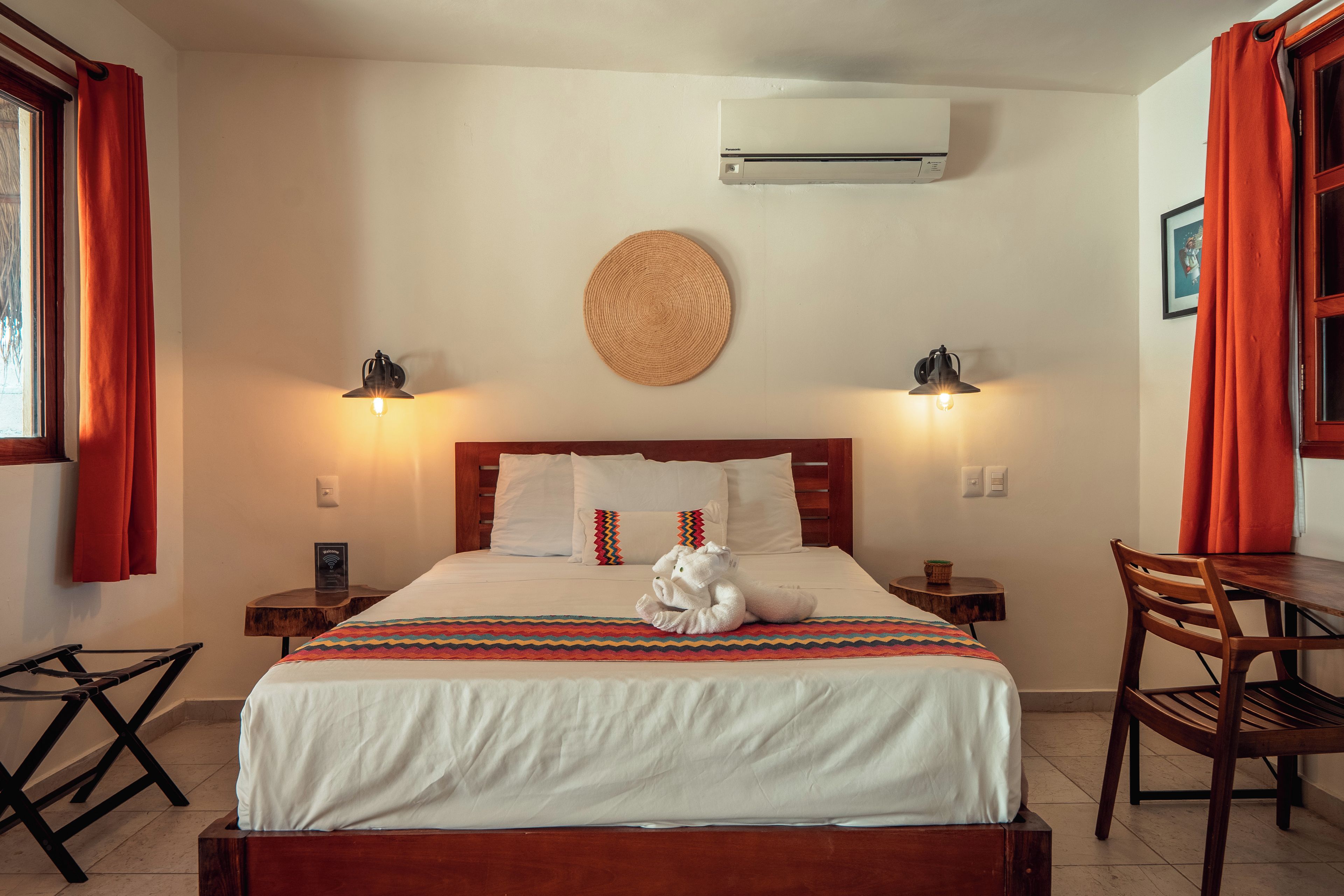 Habitación de hotel con cama, dos lámparas de noche, aire acondicionado de pared y una pequeña mesa con silla.