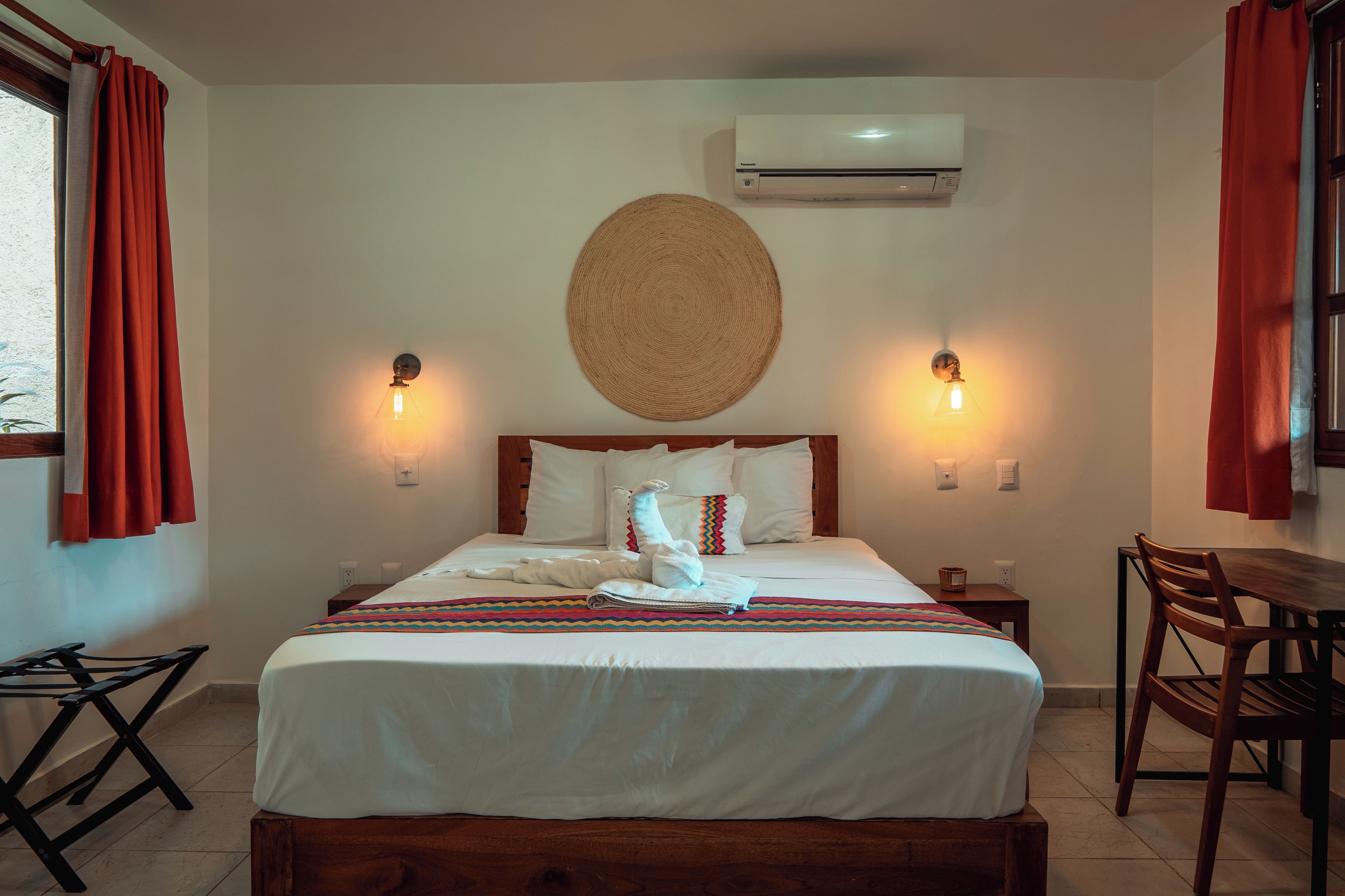 Una habitación de hotel con cama, dos lámparas de noche, aire acondicionado de pared y una mesa de comedor con sillas.