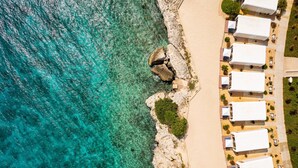 Una vista aerea di un hotel sulla costa, con un litorale roccioso, acque cristalline turchesi e diverse tende bianche disposte lungo la spiaggia.