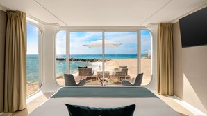 Camera d'hotel con un letto grande, vista sulla spiaggia e un tavolo con sedie davanti alla finestra.