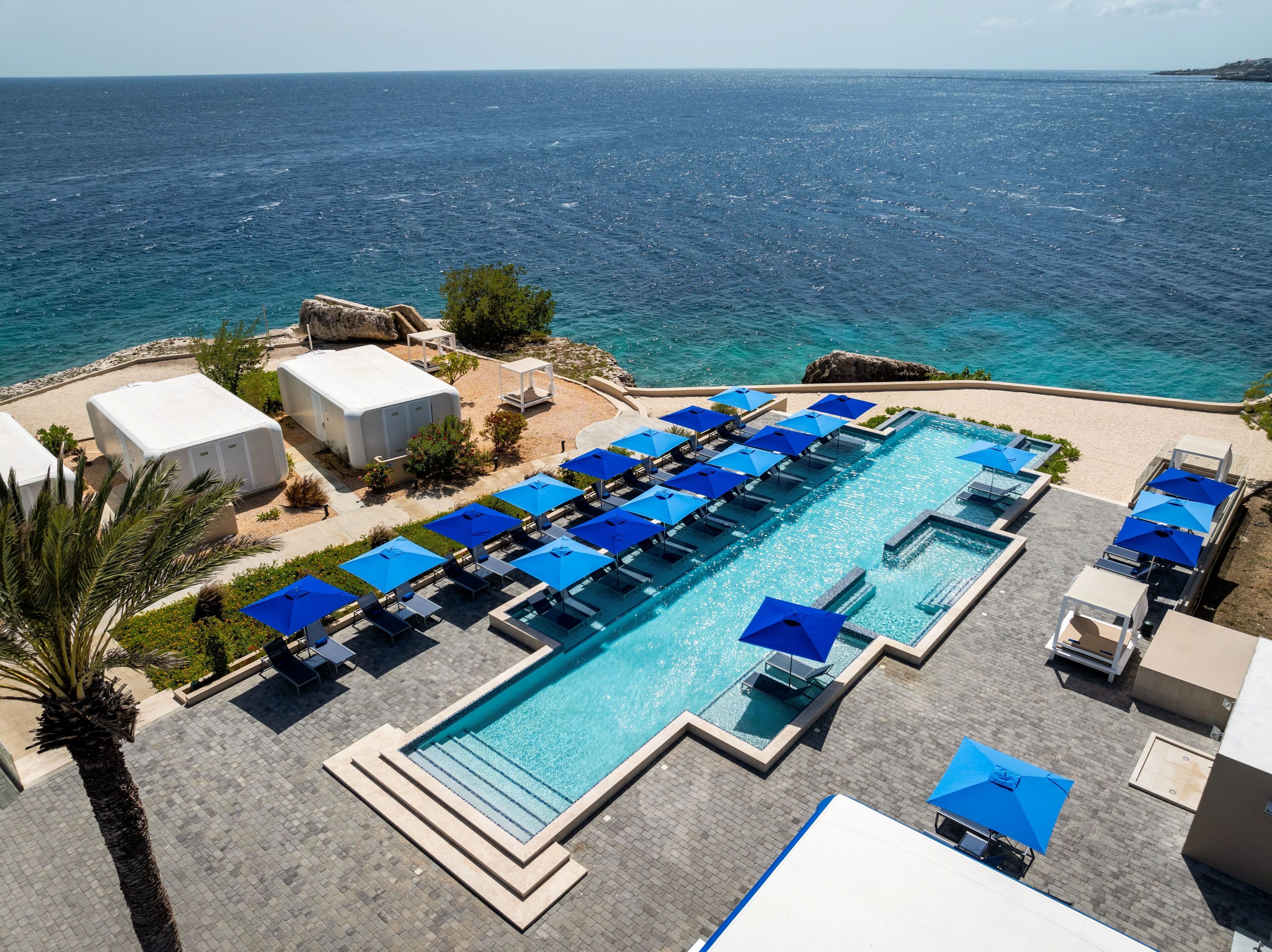 A large rectangular pool with multiple blue umbrellas and lounge chairs, surrounded by a paved deck and a view of the sea.