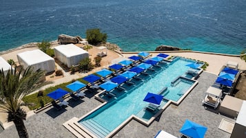 A large rectangular pool with multiple blue umbrellas and lounge chairs, surrounded by a paved deck and a view of the sea.