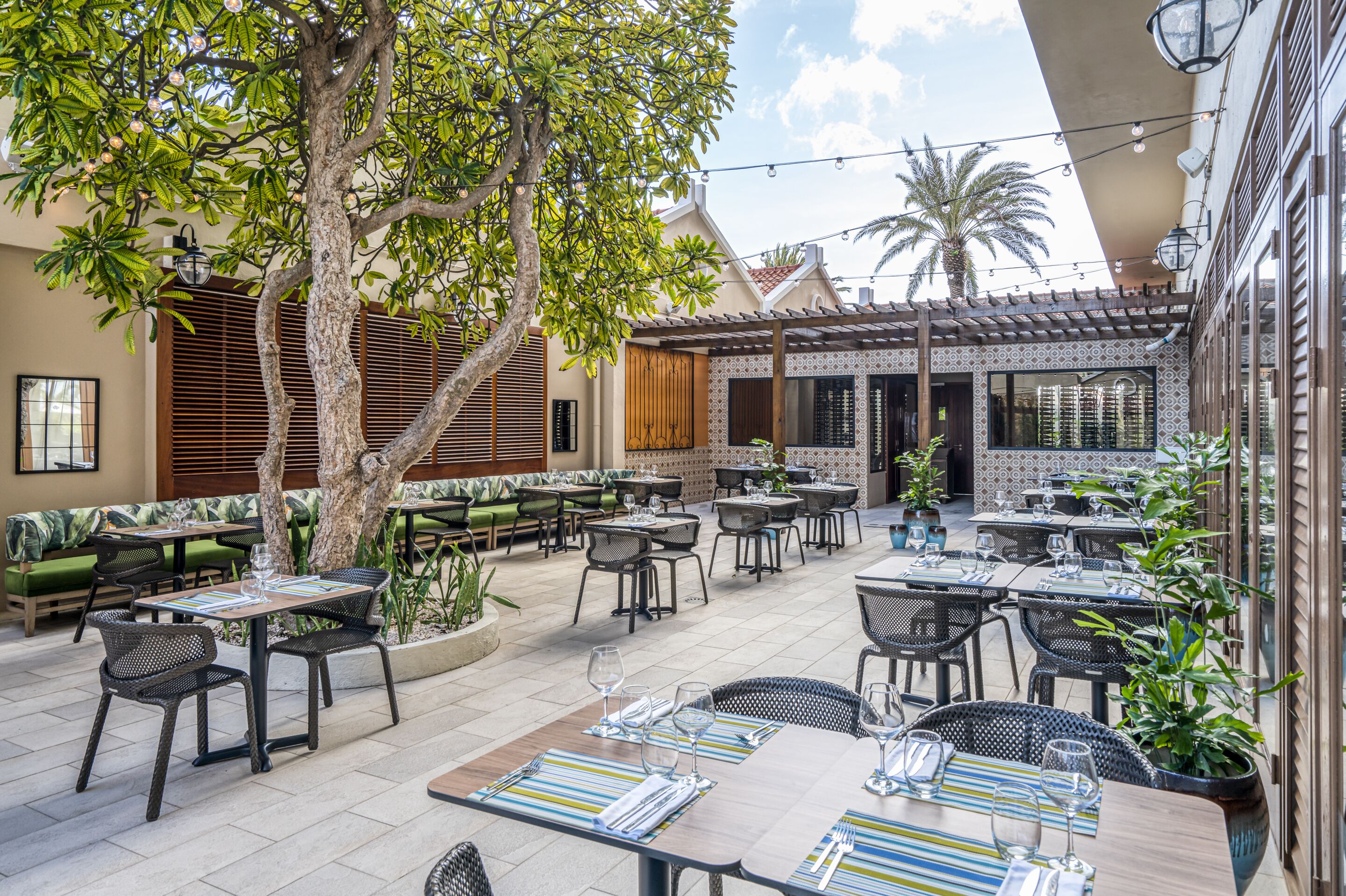 Un patio al aire libre con mesas y sillas, rodeado de follaje y detalles arquitectónicos.