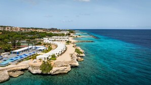 Un hotel sulla costa con una fila di cabine bianche lungo la spiaggia, acque turchesi trasparenti e vista sulla città sullo sfondo.