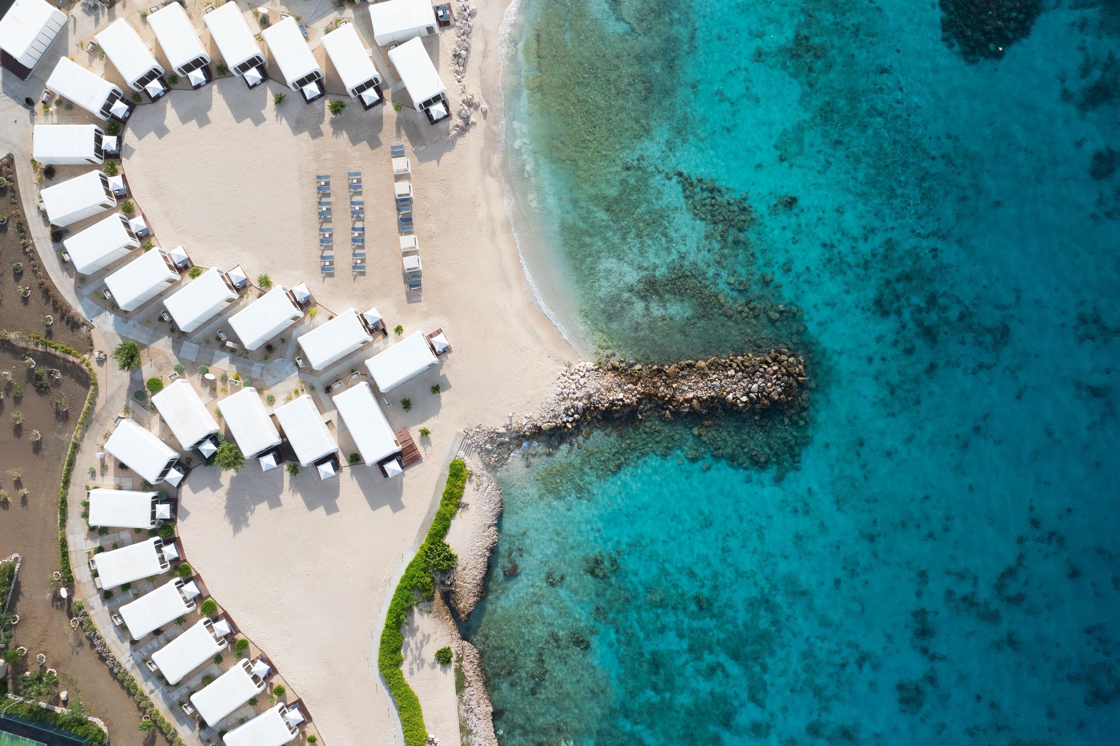 An aerial view of a beachside resort with rows of white tents and a rocky shoreline.