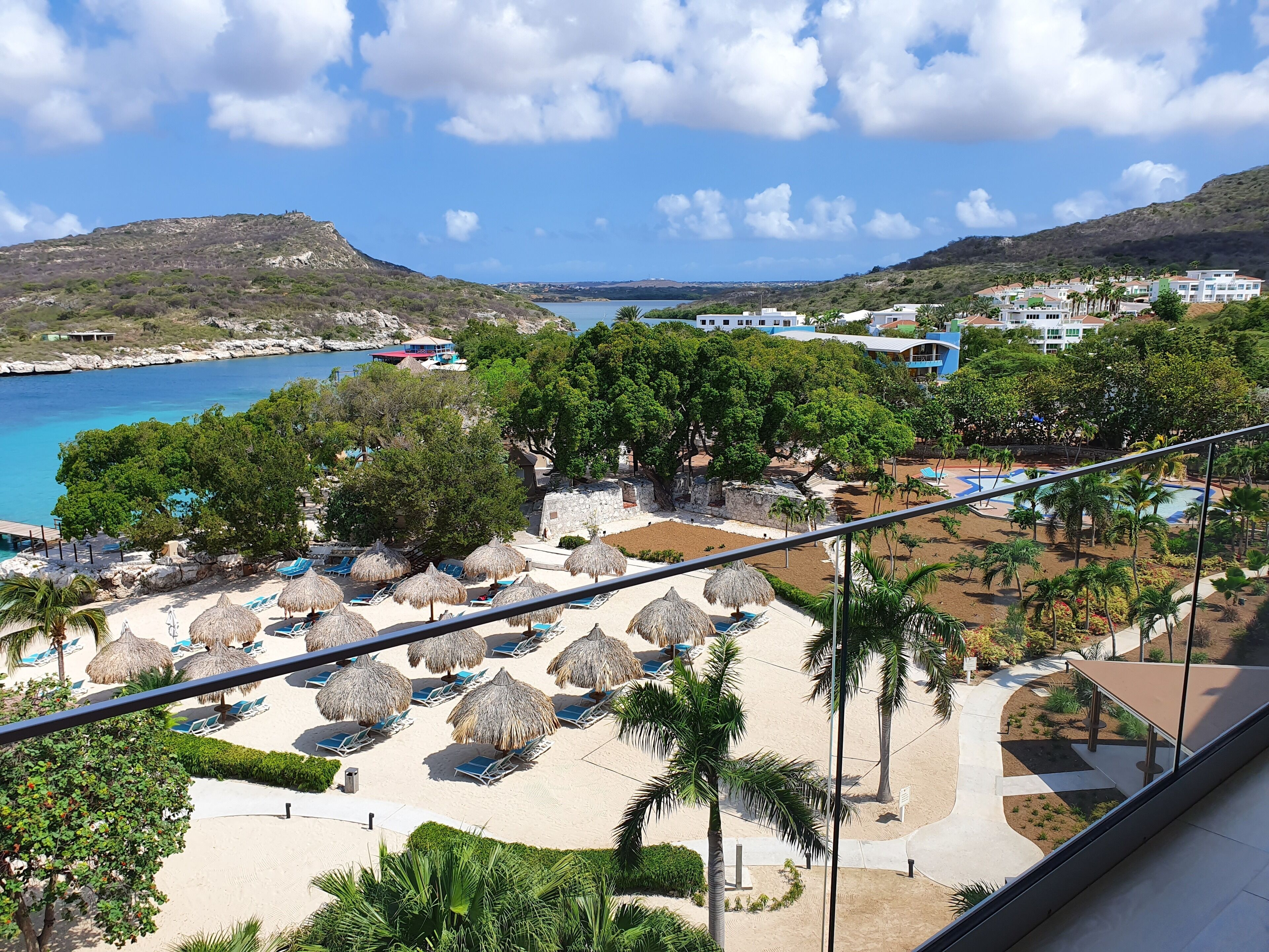 Een resort met een strandgedeelte, ligbedden en rieten parasols, een zwembad en uitzicht op de zee en heuvels.